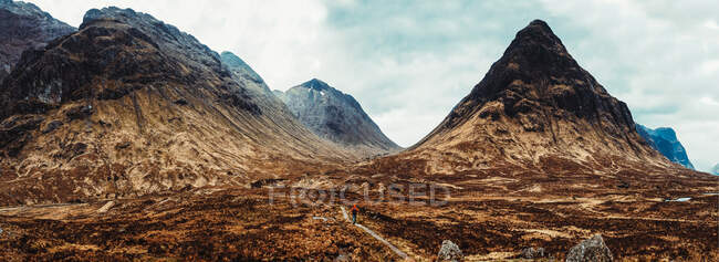 Personne méconnaissable portant un manteau rouge marchant à travers les montagnes pittoresques de l'Écosse — Photo de stock