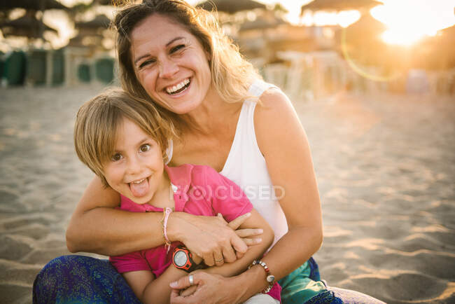 Mulher rindo bonita abraçando menino bonito mostrando língua enquanto sentados juntos na praia em sol brilhante — Fotografia de Stock