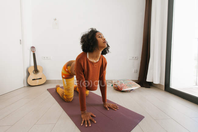 Afro-américaine jeune femme effectuant pose de yoga avec les yeux fermés sur un tapis à la maison — Photo de stock
