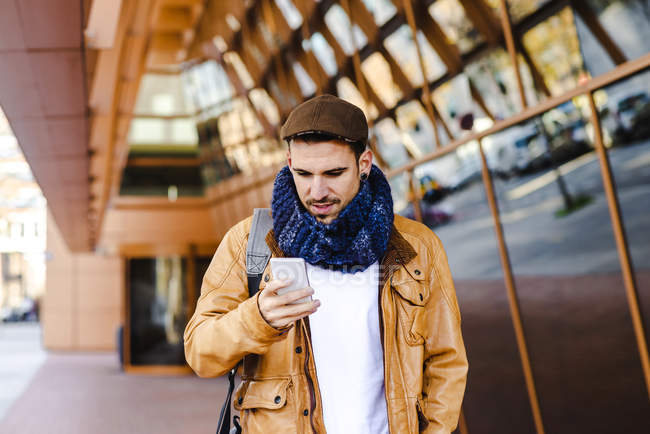Модный мужчина в повседневной одежде с помощью мобильного телефона во время прогулки возле стеклянной стены современного здания — стоковое фото