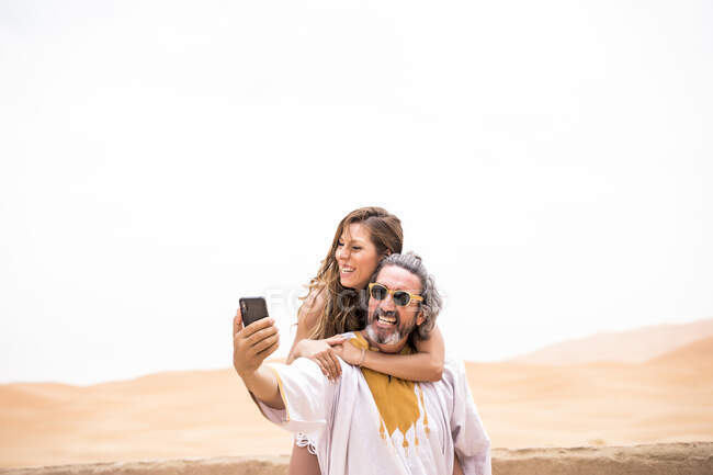 Middle-aged man with woman piggyback taking selfie expressively on terrace against sandy desert, Morocco — Stock Photo