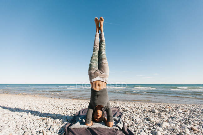 Joven afroamericana en postura de cabecera de yoga sobre fondo de mar tranquilo en día soleado - foto de stock