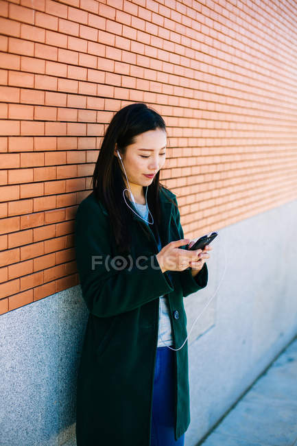 Jovem mulher asiática ouvindo música e navegando smartphone enquanto se inclina na parede de tijolos na rua da cidade — Fotografia de Stock