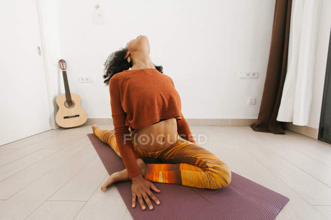 Афроамериканка, сидящая в позе йоги на коврике в светлой комнате — стоковое фото
