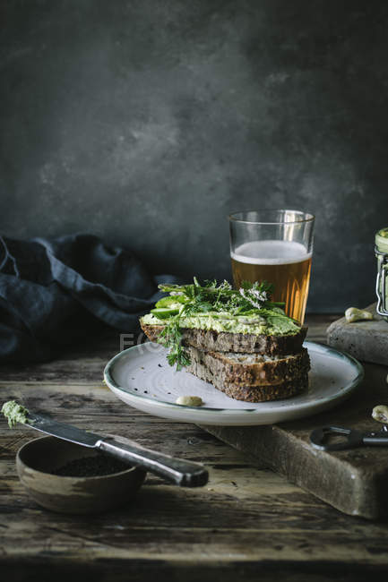 Tostadas con paté de anacardo verde, hierbas y rodajas de pepino con vaso de cerveza sobre tabla de madera - foto de stock