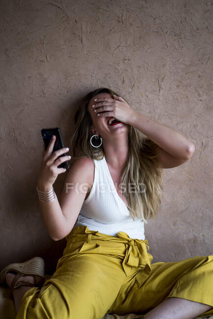 Mulher loira feliz sentada no sofá no terraço em estilo oriental e usando um telefone celular em Marrocos — Fotografia de Stock