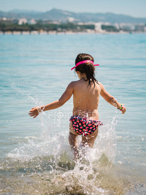 Vista trasera de chica linda alegre anónima jugando en el agua del mar en el fondo de la orilla del mar tranquila - foto de stock