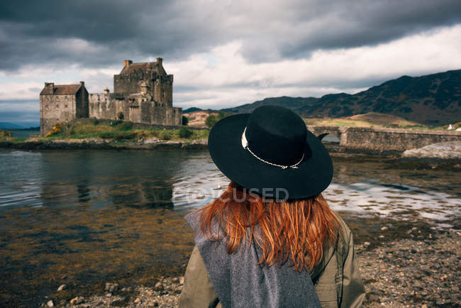 Back view of stylish woman wearing a hat contemplating old stone castle on coast in mountains, Scotland — Stock Photo