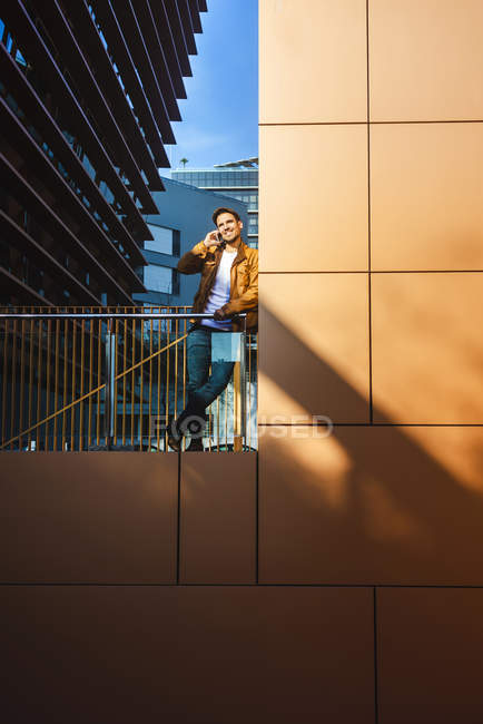 Homme positif en tenue élégante parlant sur téléphone portable tout en se tenant debout dans un balcon en verre moderne sur le bâtiment contemporain le jour ensoleillé — Photo de stock