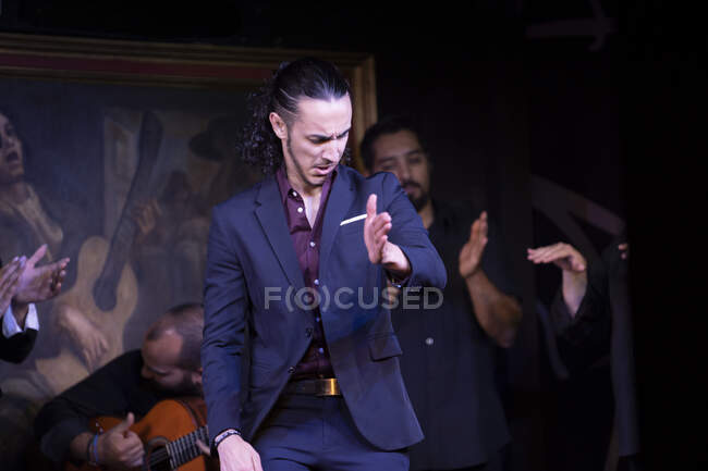 Uomo in costume blu danza flamenco vicino a musicisti maschi ispanici durante la performance contro la pittura sul palco scuro — Foto stock