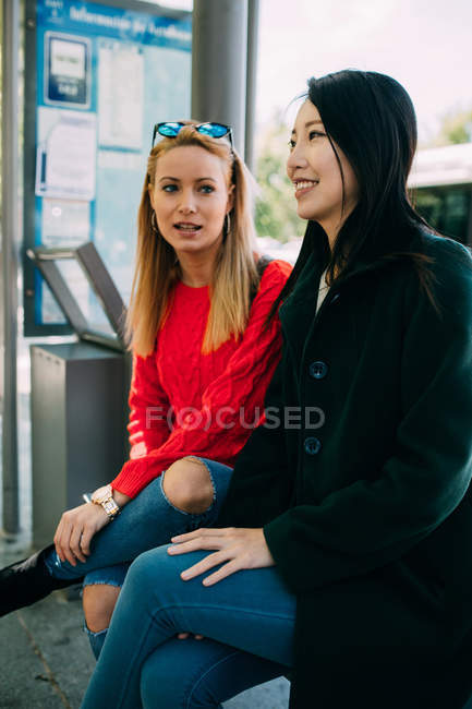 Joven mujer asiática y amigo caucásico sentado en el banco y esperando el autobús juntos - foto de stock