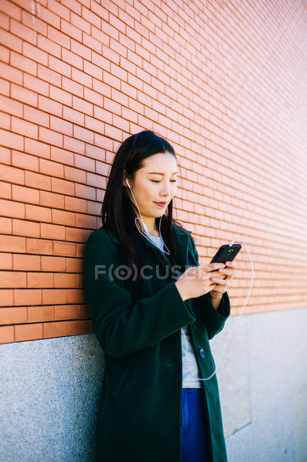 Young Asian woman listening to music and browsing smartphone while leaning on brick wall on city street — Stock Photo