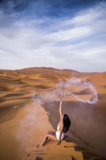 Cheerful stylish brunette holding arm raised with burning smoky firework sitting in desert of Morocco — Stock Photo