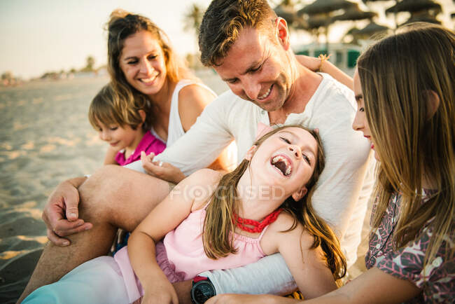 Adulto amante hombre y mujer con alegre hijo e hijas sentados juntos en la playa en la espalda iluminada - foto de stock