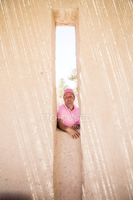 Толстый мужчина средних лет в розовой шляпе и рубашке в поло смотрит в сторону, выглядывая из узкого окна в каменной стене в солнечный день — стоковое фото