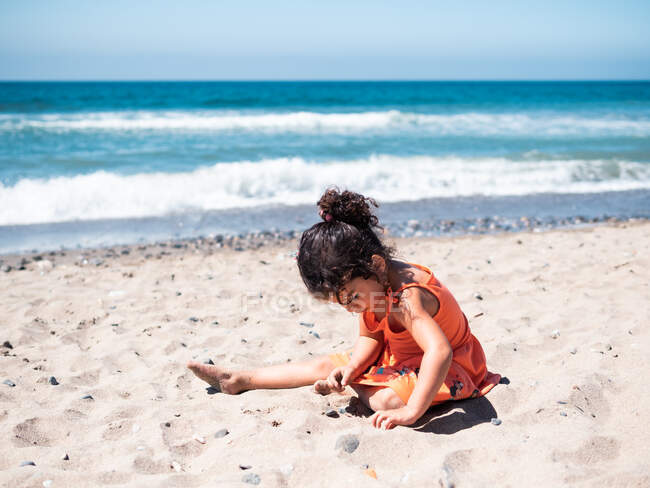 Back view of female kid in orange dress sitting on sandy seaside and playing — Stock Photo