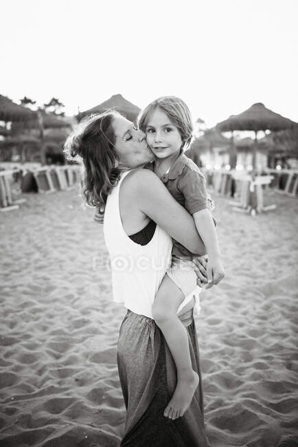 Mutter umarmt und küsst niedlichen Jungen, während sie zusammen am Strand bei strahlendem Sonnenschein steht, Schwarz-Weiß-Foto — Stockfoto