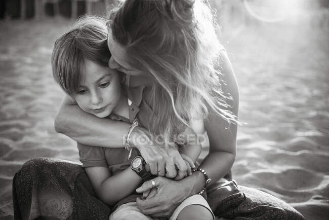 Mãe abraçando e beijando menino bonito enquanto sentados juntos na praia em sol brilhante — Fotografia de Stock