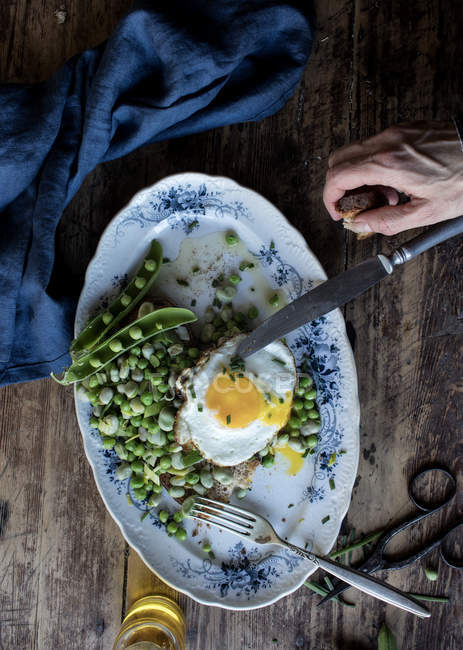 Дегустация человеческой руки из подаваемой тарелки с зеленым горошком и жареным яйцом на деревянном столе — стоковое фото
