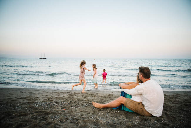 Homme et femme aimant les adultes assis à la plage tandis que leurs fils et leurs filles jouent ensemble à côté de la mer — Photo de stock