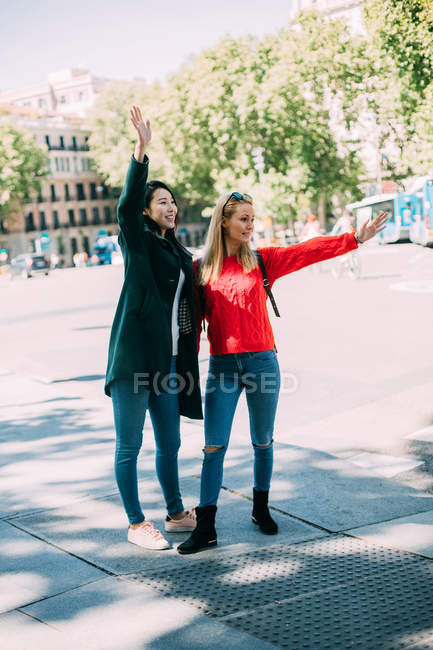 Щасливі багаторасові дами в стильних вбраннях посміхаються і махають руками, стоячи на вулиці міста в сонячний день разом — стокове фото