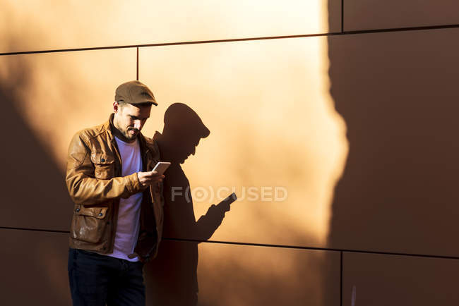 Positiver Mann in stylischem Outfit mit Handy, während er sich an sonnigem Tag an die Wand lehnt — Stockfoto
