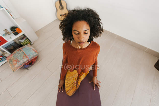 Cara de mujer joven afroamericana sentada en postura de yoga con los ojos cerrados en la estera en la sala de luz - foto de stock