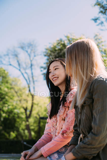 Dos mujeres jóvenes multirraciales en trajes casuales hablando y mirándose mientras están sentadas en el banco en el parque - foto de stock