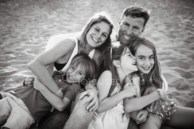 Adult loving man and woman with son and daughters sitting together on beach in back lit smiling at camera — Stock Photo