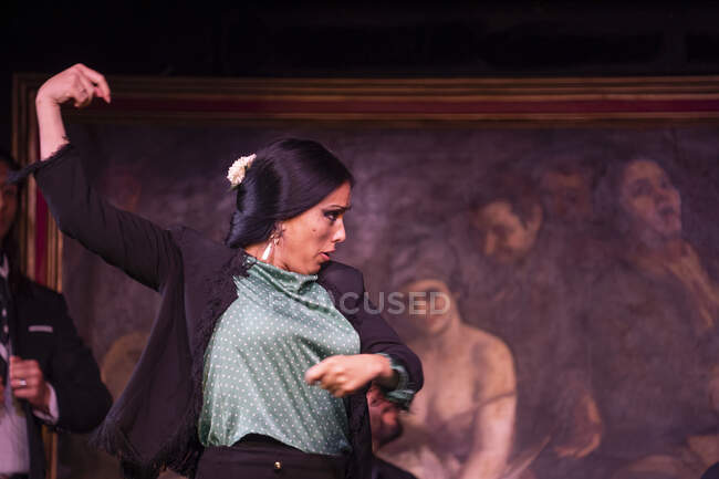Femme en costume noir dansant le flamenco près des musiciens hispaniques masculins pendant la représentation contre la peinture sur scène sombre — Photo de stock