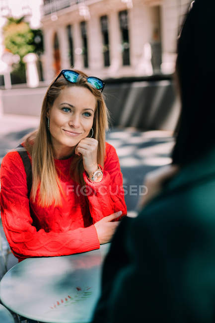 Bela jovem fêmea de malha camisola vermelha sorrindo e ouvindo amigo enquanto sentado à mesa no café ao ar livre juntos — Fotografia de Stock