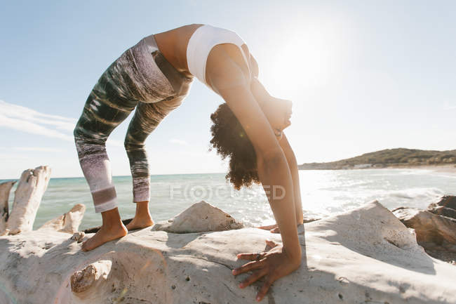 Afrikanerin steht bei sonnigem Wetter in Yoga-Haltung auf einer Brücke vor dem Hintergrund der ruhigen See — Stockfoto