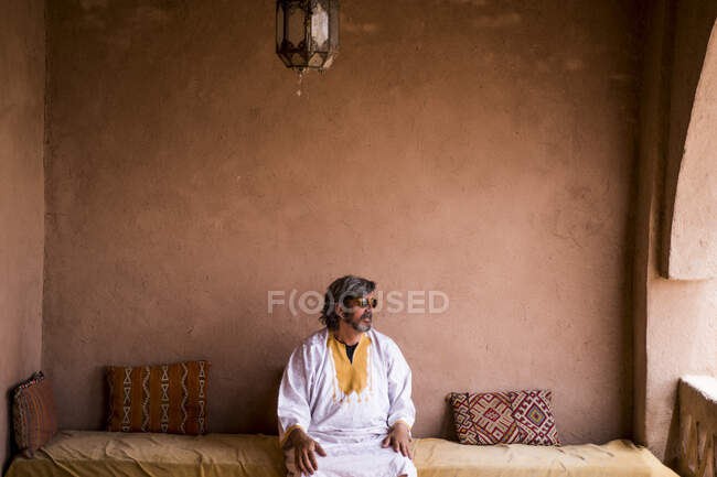 Homme adulte en vêtements longs assis sur le canapé sur la terrasse avec clôture en pierre de style oriental, Maroc — Photo de stock