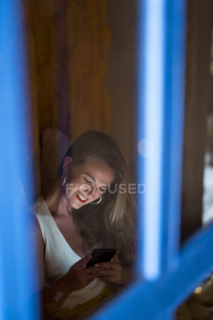 Young lady smiling and using smartphone while sitting on soft bed in dark room in Morocco — Stock Photo