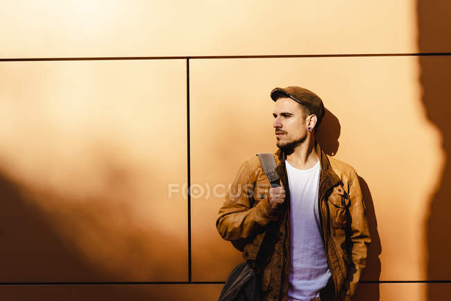 Молодий чоловік в стильному вбранні, дивлячись далеко, спираючись на стіну на вулиці в сонячний день — стокове фото