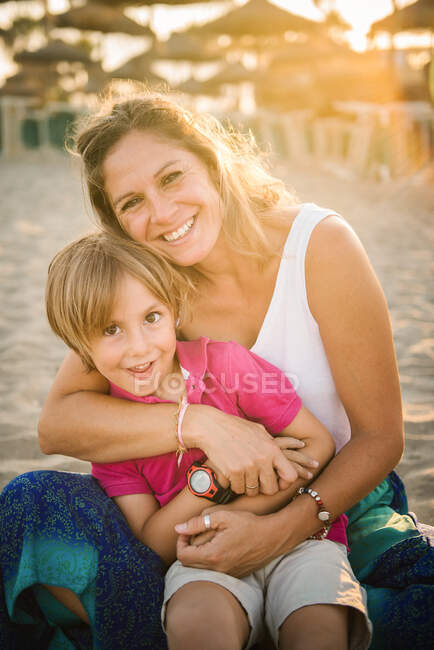 Beautiful laughing woman embracing cute cheerful boy while sitting together on beach in bright sunshine — Stock Photo