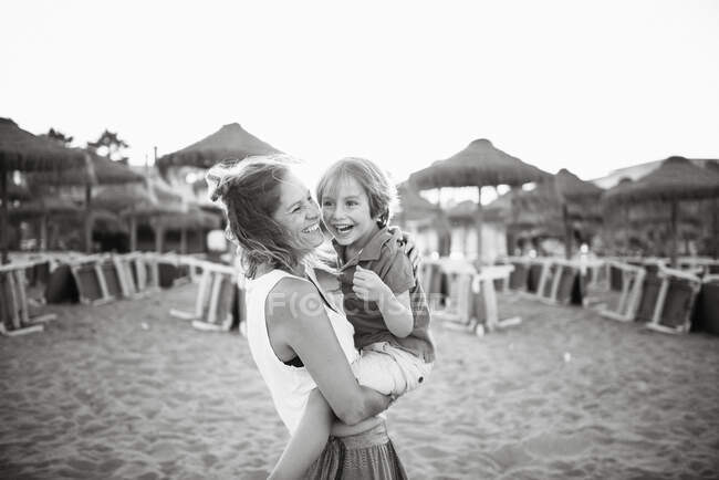 Вид сбоку смеющейся женщины с веселым игривым сыном на руках, стоя на пляже на закате, черно-белое фото — стоковое фото