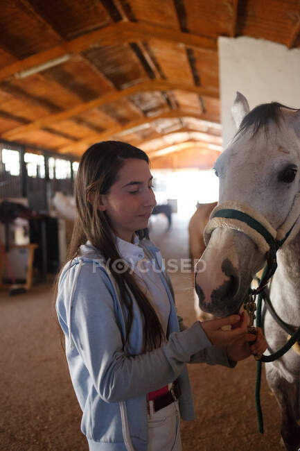 Teenage girl embracing with small pony in cute hat on ears standing inside of stable — Stock Photo