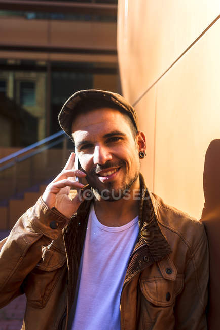 Positiver Mann in stylischem Outfit, der an sonnigem Tag an der Wand lehnt und mit dem Handy telefoniert — Stockfoto