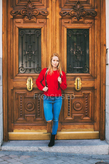 Jovem mulher positiva em roupa elegante olhando para a câmera enquanto se inclina na porta ornamental do antigo edifício na rua da cidade — Fotografia de Stock