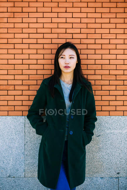 Pensativo mujer asiática en traje de moda y mirando hacia otro lado mientras se apoya en la pared de ladrillo - foto de stock