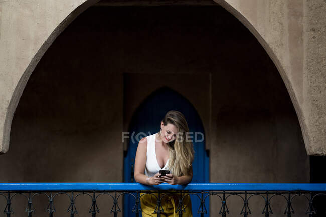 Веселая красивая женщина в стильном наряде улыбается и просматривает смартфон, опираясь на балконные перила старинного здания в Марокко — стоковое фото