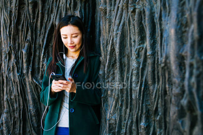 Schöne asiatische Frau in stilvollem Outfit, Musik hörend und Smartphone benutzend, während sie sich an raue Wand mit Relief von Baumwurzeln lehnt — Stockfoto