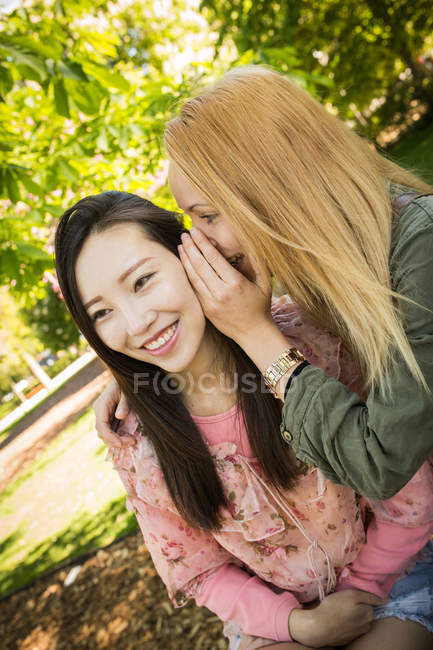 Молода кавказька жінка посміхається і шепоче таємницю до вуха усміхаючись азіатській подрузі, проводячи час у парку разом — стокове фото