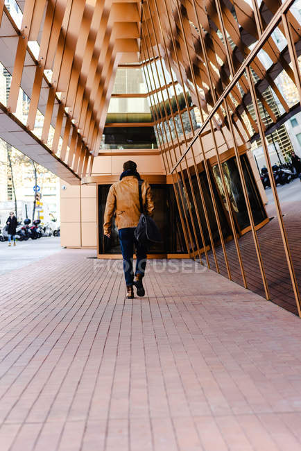 Обратный вид на неузнаваемого модного мужчину в повседневной одежде, идущего рядом со стеклянной стеной современного здания — стоковое фото