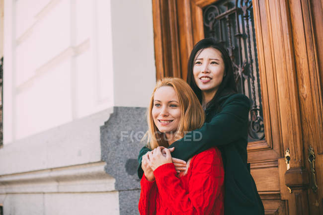 Happy Asian woman smiling and looking away while standing against ornamental door of aged building and embracing Caucasian friend — Stock Photo