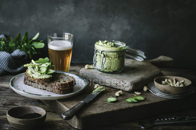 Tostadas con paté de anacardo verde, hierbas y rodajas de pepino con tarro y vaso de cerveza sobre tabla de madera - foto de stock