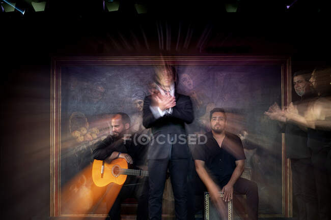 Man in black costume dancing flamenco near Hispanic male musicians during performance against painting on dark stage — Stock Photo