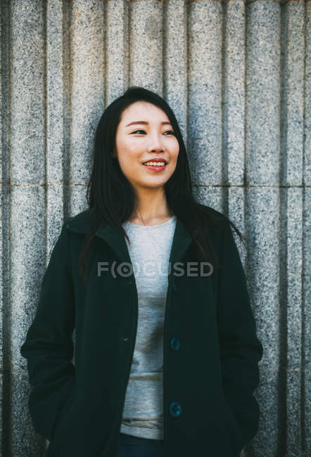 Young Asian woman in elegant coat smiling and looking at camera while leaning on marble wall on city street — Stock Photo