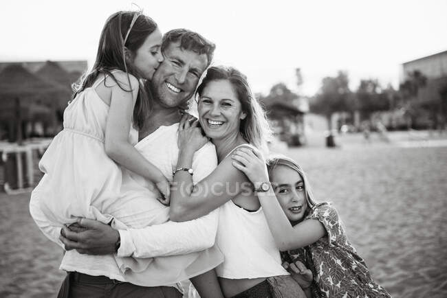 Adult loving man and woman with daughters standing together on beach in back lit smiling at camera, black and white photo — Stock Photo